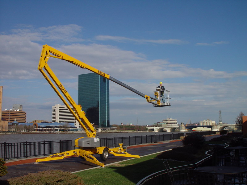 Żółty podnośnik platformowy Bil-Jax 5533A z operatorem w kasku, na tle miejskiego krajobrazu z rzeką i wieżowcem.